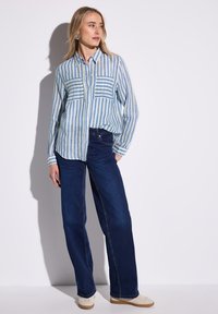 Camicia a righe blu chiaro e bianca, con due tasche sul petto, abbinata a jeans a gamba larga blu scuro e scarpe beige.