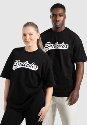 Schwarze Baumwoll-T-Shirts mit Oversized-Fit, versehen mit einem weißen "Smilodox Athlete"-Logo in Kursivschrift auf der Vorderseite.