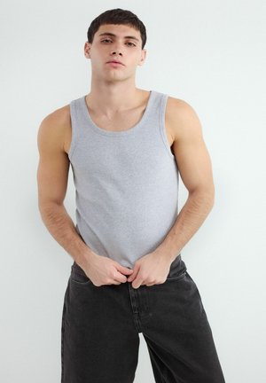 Mladý muž v sivom tielku a čiernych džínsoch stojí pred jednoduchým bielym pozadím a upravuje si nohavice.