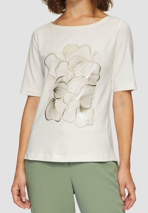 Žena v krémovém topu s krátkým rukávem a zlatobéžovým abstraktním květinovým vzorem, který doplnila lehce zelenými kalhotami.