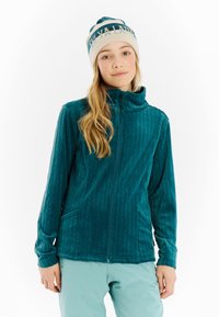 Jongere in een teal ritssluiting fleecejack, lichtblauwe broek en beige beanie met teal tekst, staande tegen een witte achtergrond.