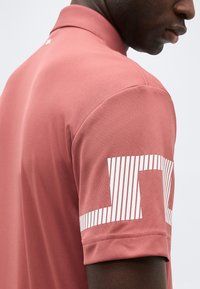 Άνδρας που φοράει μια μπλουζα πόλο κοντομάνικη σε απαλό ροζ χρώμα με γραφικό κείμενο με λευκές λωρίδες στο πάνω μέρος του μανικιού, εμφανιζόμενος από τον ώμο μέχρι το μέσο του προσώπου.