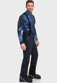 Mann trägt eine blau gemusterte Jacke, schwarze Skihosen mit Trägern und schwarze Skischuhe, vor einem einfarbigen Hintergrund stehend.