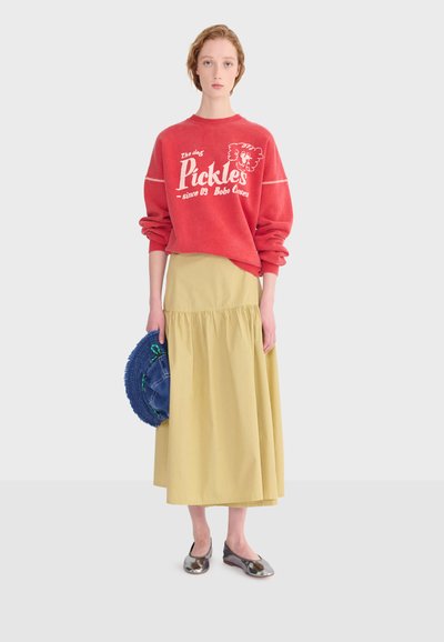 Personne portant un sweat-shirt rouge avec le texte « Pickles », une jupe midi jaune, des ballerines argentées, tenant un chapeau tissé bleu sur un fond uni.