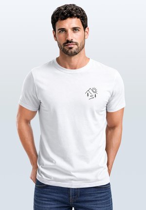 Mann mit kurzen dunklen lockigen Haaren und Bart, der ein weißes T-Shirt mit einer kleinen schwarzen Grafik von Berg und Sonne auf der Brust trägt.