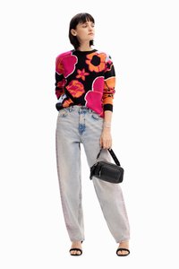 Maglione lavorato a maglia con audaci motivi floreali nei colori rosa, arancione e viola; abbinato a pantaloni in denim baggy di colore blu chiaro e una piccola borsa a tracolla nera.