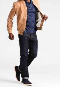 Veste bomber en daim beige sur une chemise polo bleu marine à pois, jean en denim foncé, et baskets bleu marine à semelles claires avec les poignets retroussés.