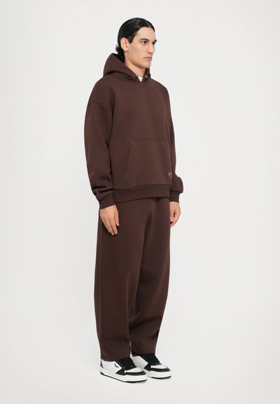 Hanorac maro cu o buzunar frontal, asortat cu pantaloni de trening maro, largi. Sneakers albi cu negru completează ținuta. Textură netedă.