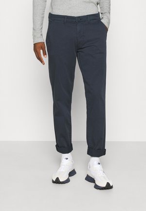 Mørkeblå chinos med lige pasform, optrillede ærmer og sidelommer, parret med hvide sneakers og en grå langærmet skjorte.