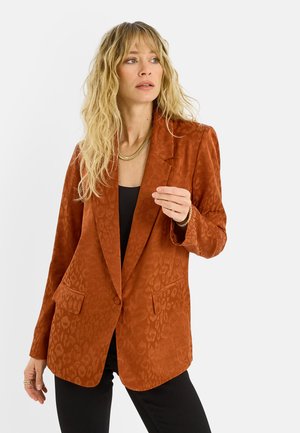 Mujer rubia que lleva una blazer estampada color óxido sobre una camiseta negra y pantalones negros, posando frente a un fondo blanco liso.