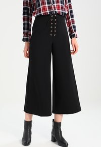 Svarta vida byxor med snörning framtill som har metallöglor. Slät tyg, kompletterad med en rutig skjorta och svarta ankelboots.