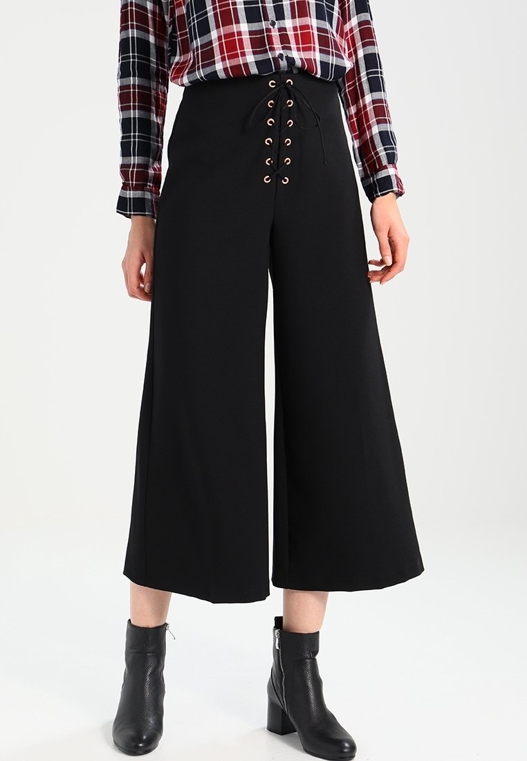 Svarta vida byxor med snörning framtill som har metallöglor. Slät tyg, kompletterad med en rutig skjorta och svarta ankelboots.