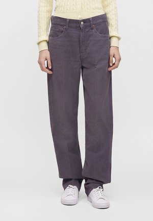 Personne portant un pantalon large en velours côtelé gris foncé, un pull en maille crème et des baskets blanches, debout sur un fond uni.