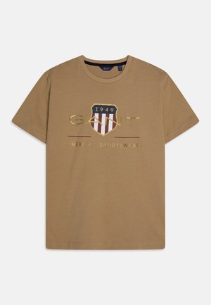 Jasnobrązowy T-shirt z krótkim rękawem i okrągłym dekoltem, z logo w kształcie tarczy z napisem "1949", "GANT" i "American Sportswear" w złotym kolorze.