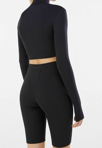 Svart träningsoverall som består av en kortärmad topp och högmidjade shorts. Slät textur med en figursydd siluett och minimal detaljering.