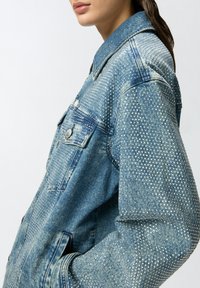 Trapera od trapera u plavoj boji, ukrašena metalnim maticama na ramenima i rukavima. Klasična ovratnik i detalj dugmadi sprijeda.