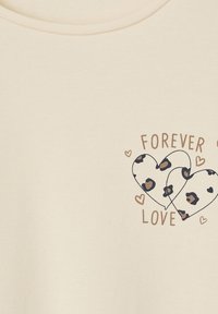 Cremfarbens T-Shirt mit Rundhalsausschnitt. Enthält zwei Herzen; eines hat ein Leopardenmuster und das andere eine einfache Kontur, mit dem Text "FÜR IMMER LIEBE."