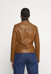 Brown leather jacket with exposed stitching, a slim fit, and a slight sheen. Zipper details and a collar complete the design. Blue denim visible below.