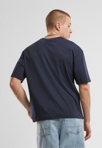 Tricou din bumbac bleumarin, cu mâneci scurte, croială lejeră, gât rotund, textură netedă, cusături vizibile, purtat cu blugi de culoare bleu deschis.