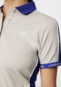 Ljusgrå polo T-shirt med blå ribbad krage och blå ärmar. Har en vit logo och kontrasterande vit piping längs sömmarna.