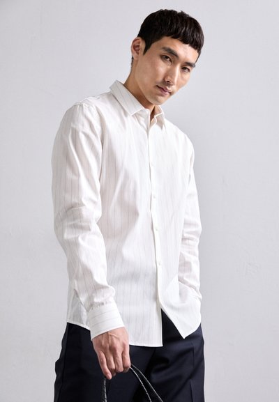 Camisa blanca de manga larga con finas rayas verticales, cuello clásico y cierre de botones, combinada con pantalones oscuros. La tela parece suave.