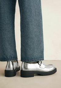 Zapatos metálicos plateados con puntera redonda, suela de goma negra y diseño minimalista, combinados con jeans de mezclilla azul holgados.