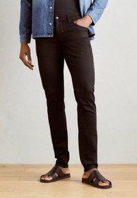 Fekete, slim fit farmernadrág, öt zsebes kialakítással és elöl gombos záródással, barna papucsokkal egy fából készült padlón.