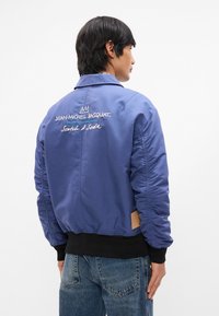 Μπλε μπουφάν με λείο, γυαλιστερό υφή. Διαθέτει ελαστικούς καρπούς, μέση και κεντημένο κείμενο στο πίσω μέρος, με δερμάτινη ετικέτα.