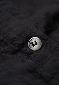 Zwart stof met een gestructureerde weving, voorzien van een ronde, zilveren knop met de opdruk "SUPERDRY" en twee knoopsgaten voor het sluiten.