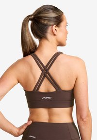 Brun sport-bh med X-back design, tillverkad av elastiskt material. Har RVRC-logotypen på bandet, slät textur och minimalistisk stil.