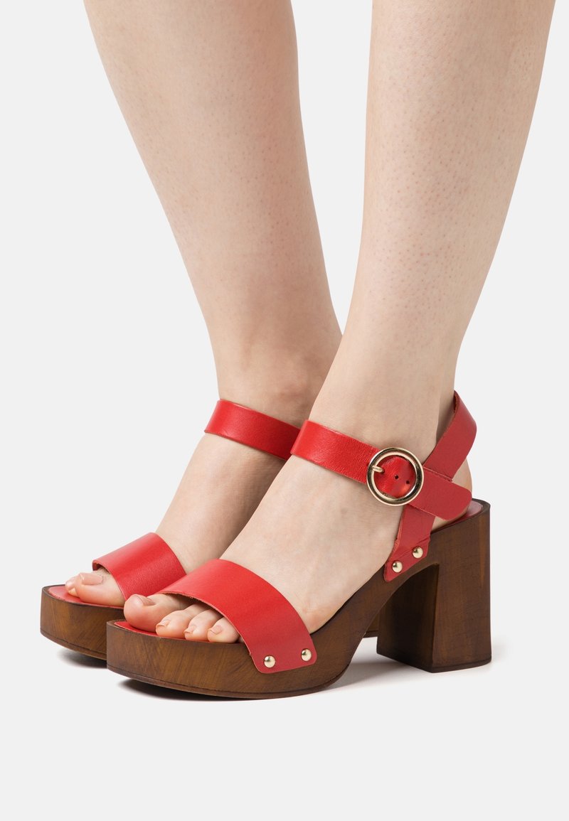 Pieds portant des sandales à brides en cuir rouge avec des talons blocs en bois hauts et une boucle dorée sur fond blanc.