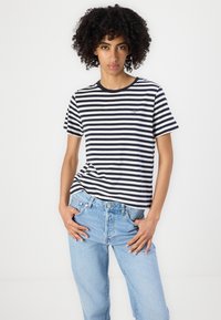 Woman with curly hair wearing black and white striped short-sleeve shirt and light blue jeans, standing with hands in pockets against plain background.