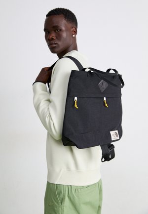 Jonge man in een licht sweatshirt en groene broek draagt een zwarte North Face-rugzak met gele ritstrekker over één schouder.