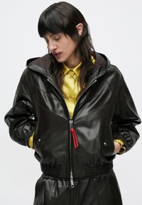 Čierna kožená bomber bunda s kapucňou, predným zipsom, elastickými manžetami a červeným sťahovacím šnúrkou. Nosená cez žltú košeľu s golierom.