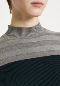 Strickrollkragenpullover in Grau und Dunkelgrün, mit horizontalen Streifen, Rippstruktur und taillierter Passform. Schlichter Hintergrund.