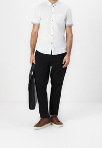 Wit shirt met korte mouwen, houten knopen en een borstzak, gecombineerd met een zwarte broek. Bruine schoenen en een zwarte tas maken de outfit compleet.