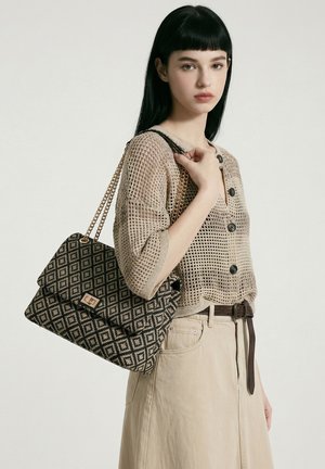 Jeune femme aux cheveux noirs portant un haut en maille beige et une jupe, portant un sac à main noir et beige à motifs avec des anses en chaîne dorée.