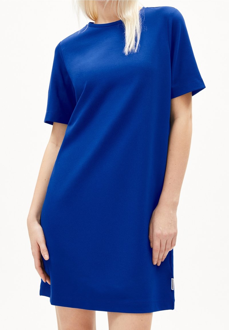 Femme portant une robe bleue royale unie, à manches courtes, longueur au genou, avec un col rond, sur fond blanc.