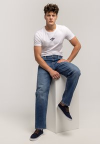 Biała bawełniana koszulka z małym granatowym logo, niebieskie jeansy dżinsowe oraz granatowe trampki z brązowymi sznurowadłami, siedzący na jednolitym tle.