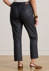 Γυναίκα με μαύρο παντελόνι ευθείας γραμμής, μπεζ τοπ, καφέ πέδιλα με λουράκι πίσω και χοντρά βραχιόλια, στέκεται σε ξύλινο πάτωμα.