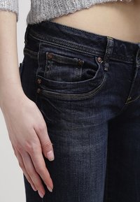 Jeans de mezclilla oscura con un diseño ajustado, que cuentan con remaches de cobre, costuras sutiles y un diseño de cinco bolsillos. Textura suave con un leve desgaste.