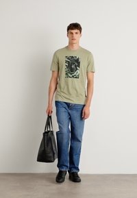Oliven grønn t-skjorte med svart og hvitt grafisk trykk, kombinert med blå denimbukser og svarte sko. Modellen bærer en svart totebag.