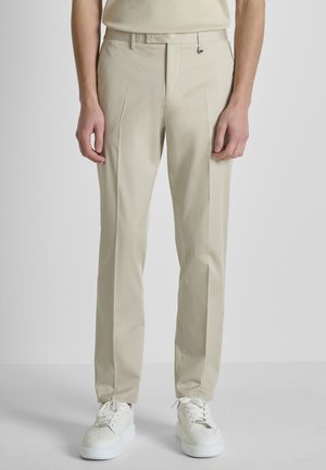 Hombre con pantalones beige a medida y zapatillas blancas de pie frente a un fondo gris claro liso.