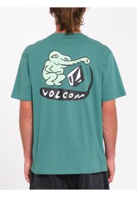 Camiseta de algodón verde con mangas cortas, que presenta un personaje de dibujos animados y el logo "Volcom" impreso en la parte posterior en blanco y negro.