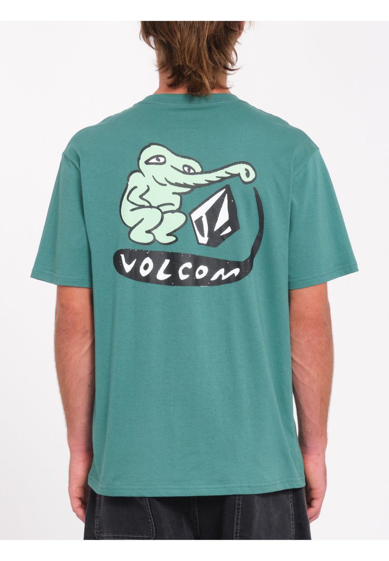 Camiseta de algodón verde con mangas cortas, que presenta un personaje de dibujos animados y el logo "Volcom" impreso en la parte posterior en blanco y negro.