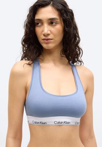Γυναίκα με σγουρά σκούρα μαλλιά που φοράει ένα ανοιχτό μπλε αθλητικό σουτιέν Calvin Klein με λευκή ελαστική ταινία με λογότυπο, στραμμένη ελαφρά προς τα αριστερά.