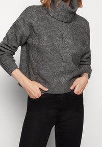 Suéter gris de punto trenzado con cuello alto, de mangas largas y puños acanalados, parcialmente metido dentro de unos jeans negros, que presenta un corte relajado y una textura suave.