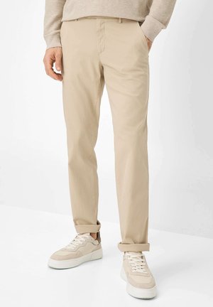 Khaki-Chinos mit geradem Schnitt, weichem Baumwollmaterial, hochgerollten Säumen und vorderen Taschen, kombiniert mit beigen Sneakers.
