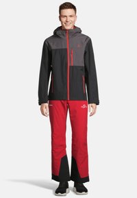 Herren wasserdichte Skijacke in Schwarz und Grau mit roten Akzenten, kombiniert mit roten Skihosen mit schwarzem Besatz und Logodetails.
