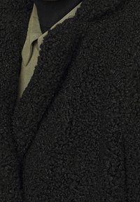 Cappotto nero texturizzato con una superficie morbida e soffice. Presenta un colletto a scialle e strati visibili sopra una camicia verde oliva sottostante.